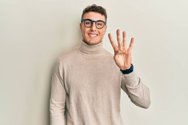 Balıkçı yaka kazak giyen İspanyol genç adam gülümseyerek kendinden emin ve mutlu bir şekilde parmaklarıyla dört numarayı gösteriyor ve işaret ediyor.. 