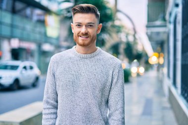 Genç beyaz adam gülümseyerek şehirde mutlu mesut duruyor..