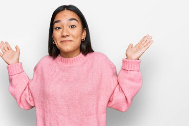 Sıradan bir kazak giyen genç Asyalı kadın elleri ve kolları havada, kafası karışık bir ifade takınıyor. şüphe kavramı. 
