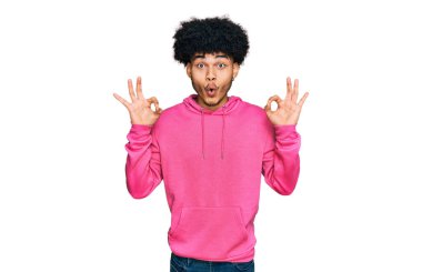 Afro saçlı, sıradan pembe kazak giyen Afro-Amerikalı genç bir adam parmaklarıyla onay sembolü yaparken şaşırmış ve şok olmuş görünüyor. çılgın ifade 