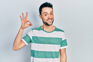 Gündelik kıyafetler giyen genç İspanyol adam gülümsüyor. El ve parmaklarıyla 