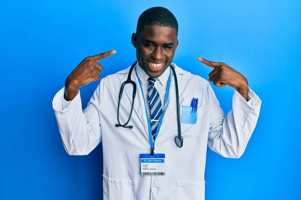 医師の制服を着た若いアフリカ系アメリカ人の男性は 陽気な笑みを浮かべて指の歯と口を指しています 歯の健康の概念 — ストック写真