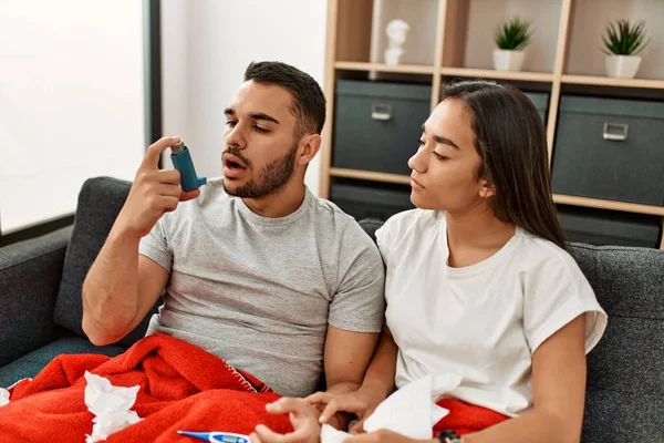 Young latin ill couple using inhaler sititng on the sofa at home.