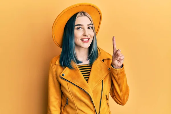 若いです現代的な女の子身に着けています黄色の帽子と革のジャケットショーと指差しますと指番号1とともに笑顔自信と幸せ — ストック写真
