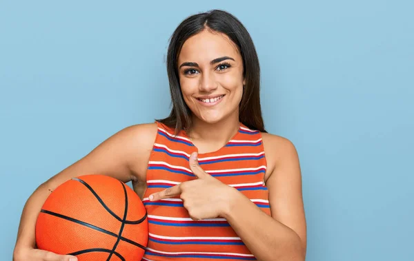 Młoda Brunetka Kobieta Trzyma Piłkę Koszykówki Uśmiechając Szczęśliwy Wskazując Ręką — Zdjęcie stockowe