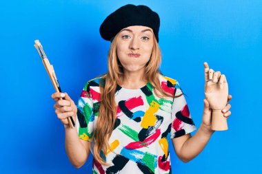 Kafkasyalı genç bir kadın sanatçı elinde küçük tahta bir manikin ve komik yüzlü fırça şişiren boya fırçaları tutuyor. Ağzı havayla şişer, havayı yakalar.. 