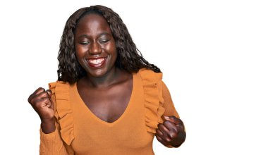 Genç Afrikalı kadın gündelik kıyafetler giyip çok mutlu ve heyecanlı bir şekilde kolları havada, gülümseyen ve başarı için çığlık atan kazanan bir jest yapıyor. kutlama konsepti. 