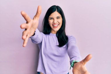 Gündelik giysiler giymiş genç İspanyol kadın kameraya bakıp sarılmak için kollarını açmış gülümsüyor. Mutluluğu kucaklayan neşeli bir ifade.. 