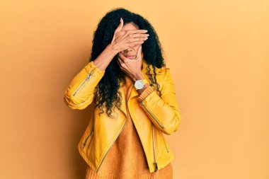 Orta yaşlı Afro-Amerikalı kadın yün kazak ve deri ceket giyip gözlerini ve ağzını elleriyle kapatıyor, şaşırmış ve şok olmuş. duygu gizleme 