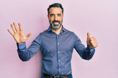 Orta yaşlı, sakallı, gri saçlı iş kıyafetleri giyen, altı numaralı parmaklarıyla işaret eden, kendinden emin ve mutlu bir adam.. 