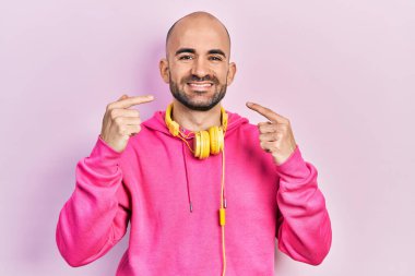 Spor kıyafetleri giyen, kulaklık takan, gülümseyen, parmaklarıyla ve ağzıyla işaret eden genç kel bir adam. diş sağlığı kavramı. 