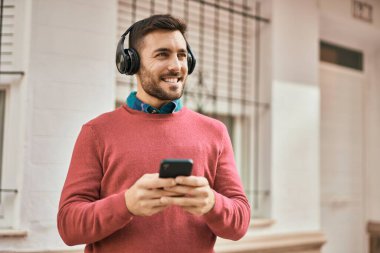 Genç İspanyol adam şehirde akıllı telefon ve kulaklık kullanarak mutlu gülümsüyor..