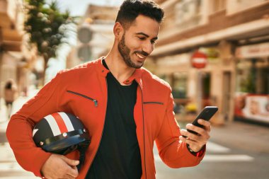 Genç İspanyol adam şehirde moto miğferi tutan akıllı telefon kullanıyor..