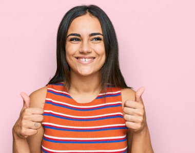 Genç esmer kadın günlük kıyafetler giyiyor başarı işareti yapıyor, elleriyle olumlu jest yapıyor, gülümsüyor ve mutlu. Neşeli ifade ve kazanan jest. 