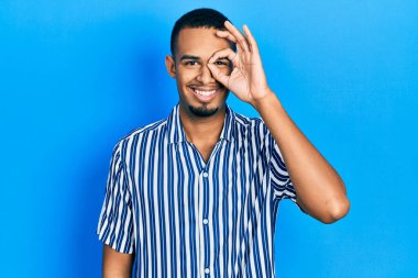 Genç Afro-Amerikan adam günlük kıyafetler giyiyor. El hareketleriyle gülümsüyor, gözleriyle mutlu yüzle parmaklarının arasından bakıyor.. 