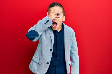 Zarif bir iş ceketi giyen küçük bir çocuk. Şoka girmiş, yüzünü ve gözlerini eliyle kapatmış, parmaklarından korkuyla bakıyor. 