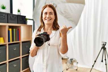 Orta yaşlı fotoğrafçı kadın fotoğraf stüdyosundaki kamerasını kullanarak baş parmağını yana kaldırıyor ve ağzı açık gülümsüyor. 