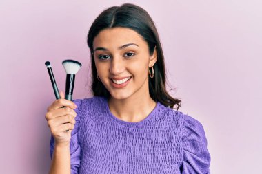 Makyaj fırçası tutan genç İspanyol kadın pozitif ve mutlu görünüyor. Duruyor ve gülümsüyor. Kendine güvenen bir gülümsemeyle dişlerini gösteriyor. 