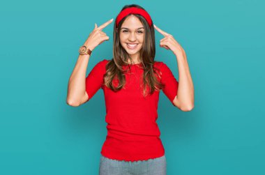 Genç esmer kadın, günlük giysiler içinde gülümseyerek iki eliyle başını işaret ediyor. Harika bir fikir ya da düşünce, iyi bir hafıza. 