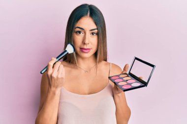 Makyaj fırçası tutan genç İspanyol kadın yüzünde ciddi bir ifadeyle rahatladı. Basit ve doğal bir şekilde kameraya bakıyorsun.. 