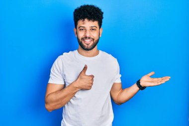 Sakallı, beyaz tişörtlü genç bir Arap. Avuç içi gösteriyor ve baş parmağıyla hareket ediyor. Mutlu ve neşeli gülümsüyor. 