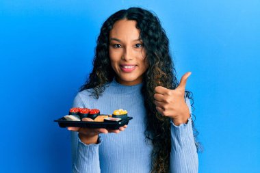 Genç Latin kadın bir tabak suşi gösteriyor mutlu ve pozitif gülümsüyor, başparmağıyla mükemmel ve onay işareti yapıyor. 