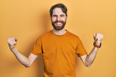 Sakallı, sıradan sarı tişörtlü beyaz bir adam çok mutlu ve heyecanlı bir şekilde kolları havada, gülümseyerek ve başarı için çığlık atarak kazanan bir jest yapıyor. kutlama konsepti. 