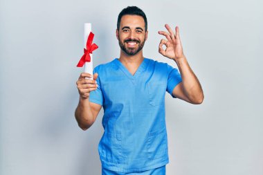 Yakışıklı, sakallı, mavi erkek hemşire üniforması giyen, diplomasını elinde tutan, parmaklarıyla imza atan, güler yüzlü, dost canlısı, mükemmel bir sembol. 