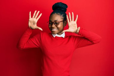 Genç Afro-Amerikalı kadın günlük kıyafetler ve gözlük takıyor ve parmaklarıyla on numarayı gösterirken kendinden emin ve mutlu gülümsüyor.. 