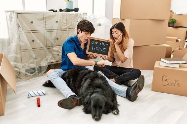 Young caucasian couple with dog holding our first home blackboard at new house looking at the watch time worried, afraid of getting late