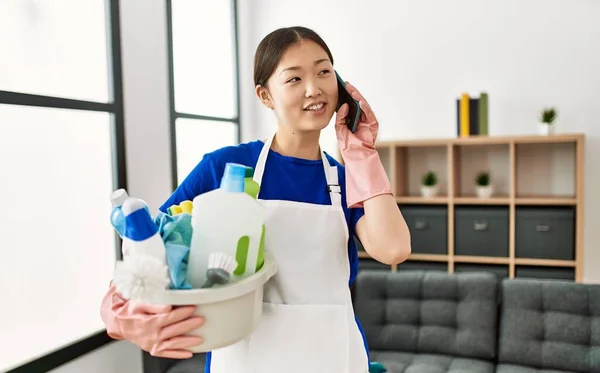 Young Chinese Housewife Holding Cleaning Products Talking Smartphone Home — Stock Photo, Image