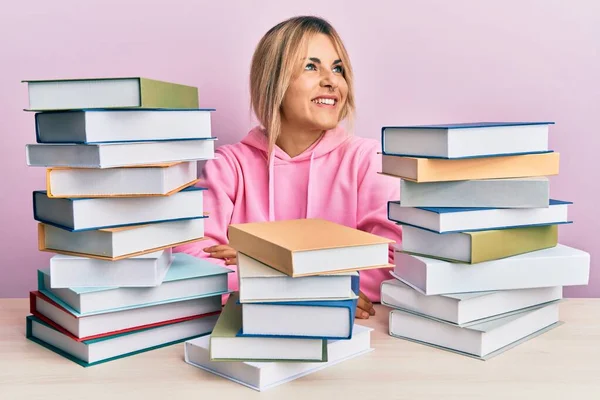 Young caucasian woman sitting on the table with books smiling looking to the side and staring away thinking.