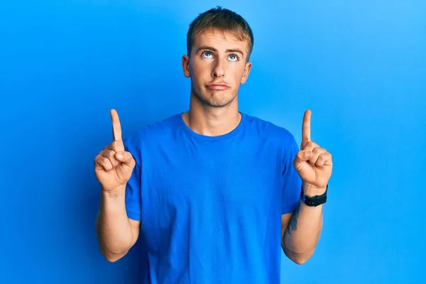 年轻的高加索人穿着休闲的蓝色T恤 面带微笑地抬起头 用手指指示方向 既不快乐又沮丧 — 图库照片