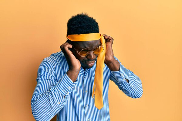 Handsome black man drunk wearing tie over head and sunglasses covering ears with fingers with annoyed expression for the noise of loud music. deaf concept.