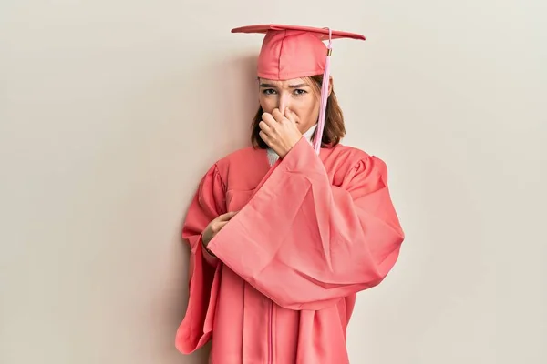 卒業キャップと式のローブを着た若い白人女性は 臭いと嫌な 耐えられない匂いを嗅ぎ 鼻の上に指で息をしています 臭いが悪い — ストック写真