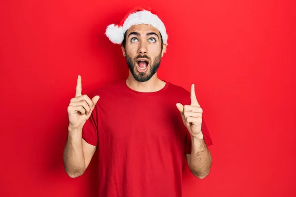 Mladý Hispánec Vánočním Kloboukem Užaslý Překvapený Když Vzhlíží Ukazuje Prsty — Stock fotografie