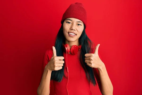 年轻的中国女人用耳机听音乐 成功的标志着她用手做了一个积极的手势 竖起大拇指 微笑着 快乐地看着 快乐的表达和胜利的姿态 — 图库照片