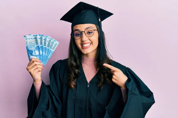 卒業制服を着た若いヒスパニック系の女性手と指で幸せなポインティング笑顔のチリペソ紙幣を保持 — ストック写真