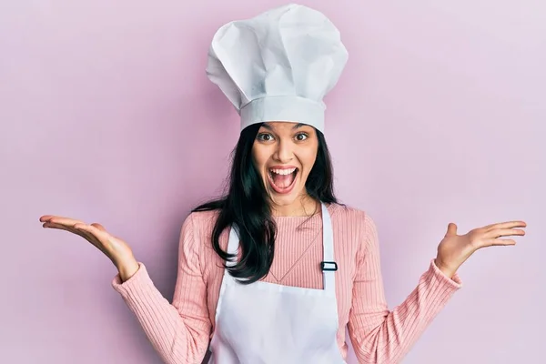 パン屋の制服を着た若いヒスパニック系の女性と幸せな笑顔で勝利を祝う帽子を調理し 手を上げて勝者の式 — ストック写真