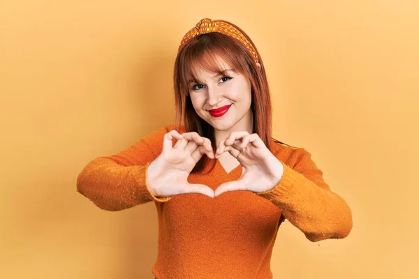 红头发的年轻女人穿着休闲的橙色毛衣 带着爱的笑容用手做心形符号 浪漫的概念 — 图库照片