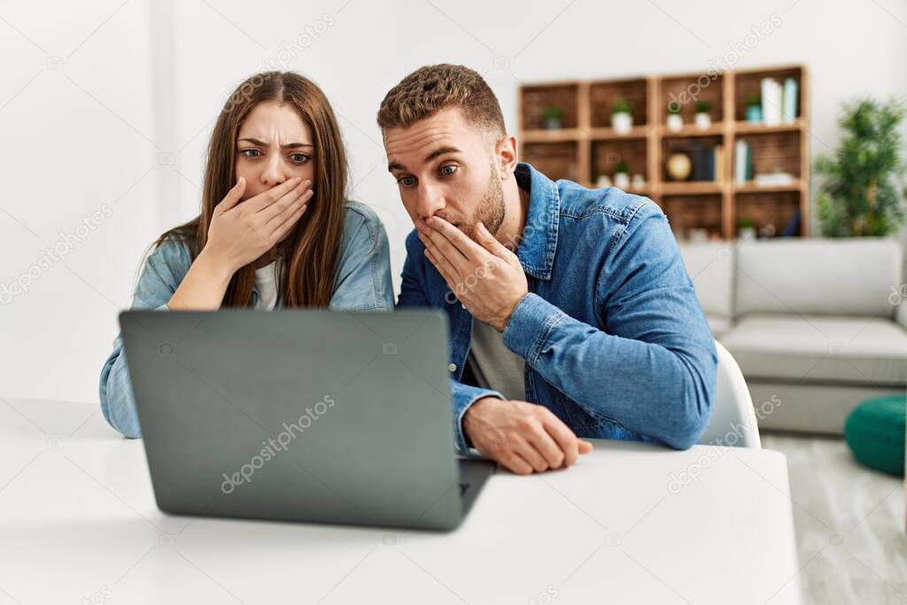 Young caucasian couple working using computer laptop at home covering mouth with hand, shocked and afraid for mistake. surprised expression 