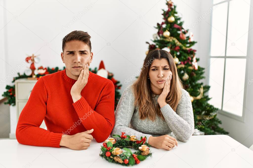 Young hispanic couple sitting at the table on christmas touching mouth with hand with painful expression because of toothache or dental illness on teeth. dentist 