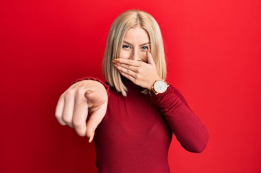 Gündelik kıyafetler giyen genç sarışın kadın sana gülüyor, eli ağzında kamerayı işaret ediyor, yüz ifadesi utanç verici. 