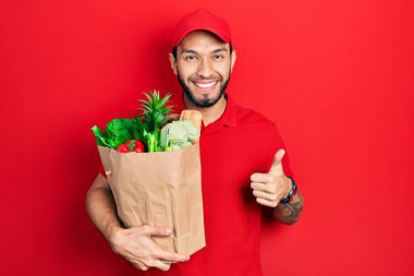 Sakallı, kurye üniformalı, marketten aldığı yiyeceklerle gülümseyen, mutlu ve olumlu gülümseyen, mükemmel ve onay işareti yapan İspanyol bir adam. 