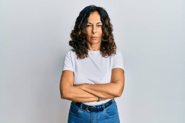 Orta yaşlı İspanyol kadın, sıradan beyaz tişört giyiyor şüpheci ve gergin, yüzünde çapraz kollu tasvip etmeyen bir ifade var. negatif kişi. 