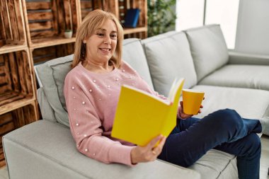 Orta yaşlı sarışın kadın gülümsüyor mutlu kitap okuyor ve evde kahve içiyor..