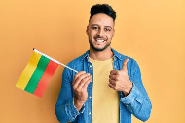 Elinde Litvanya bayrağı tutan genç Arap adam mutlu ve olumlu gülümsüyor, başparmağıyla mükemmel ve onay işareti yapıyor. 