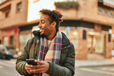 Genç Afrikalı Amerikalı adam şehirde akıllı telefon kullanarak mutlu bir şekilde gülümsüyor.