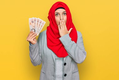 İslami tesettüre bürünmüş güzel bir İspanyol kadın elinde 5 kilo banknotla ağzını eliyle kapatıyor, şok geçiriyor ve hata yapmaktan korkuyor. şaşırmış ifade 