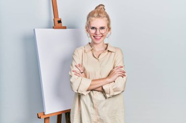 Sarı saçlı güzel beyaz kadın boyacı sehpasının yanında durup kollarını kavuşturup kameraya bakarak gülümsüyor. pozitif kişi. 